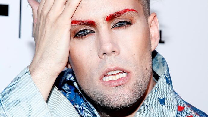 Justin Tranter, with blonde hair and red eyebrows posing in a denim jacket with a red and blue stain...