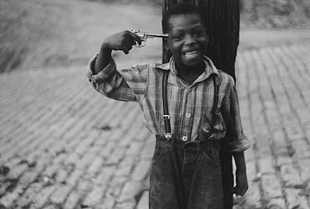 A boy wearing an overall and striped shirt smiling while holding a gun next to his temple