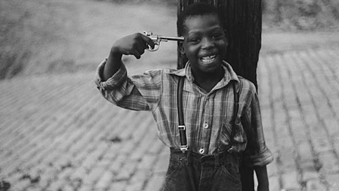 A boy wearing an overall and striped shirt smiling while holding a gun next to his temple