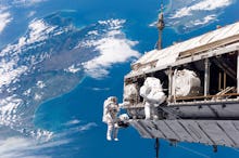 Astronauts holding on to the satellite in space with blue sky in their background.