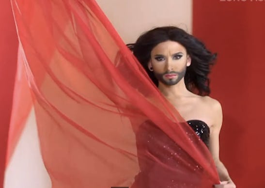Conchita Wurst in a black sequin off-the-shoulder dress holding a piece of red tulle piece of fabric