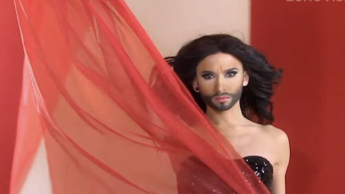 Conchita Wurst in a black sequin off-the-shoulder dress holding a piece of red tulle piece of fabric