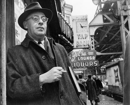A black and white photo of Saul Alinsky holding a cigarette with a liquor store in the background