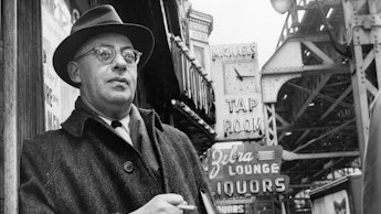 A black and white photo of Saul Alinsky holding a cigarette with a liquor store in the background