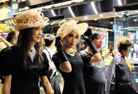 A feminine gay man in a Halloween costume with a beige sunhat and a black dress, accompanied by blac...