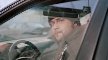 A man sitting in his car smoking a cigar while wearing a coat and a beanie