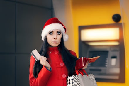 A woman in a red jacket and a christmas hat holding her credit card in front of an ATM with a confus...