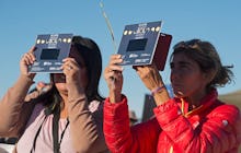 Two women standing outside using solar eclipse viewing equipment to look at the passing solar eclips...