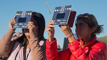 Two women standing outside using solar eclipse viewing equipment to look at the passing solar eclips...