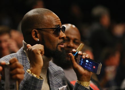R Kelly in a grey checked blazer, grey sweater with sunglasses speaking and holding his smartphone