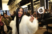 Cardi B in a white furry jacket laughing with a group of people taking a photo of her