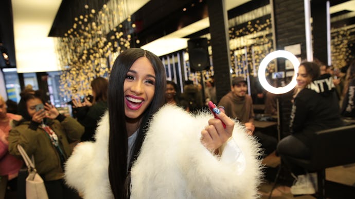 Cardi B in a white furry jacket laughing with a group of people taking a photo of her
