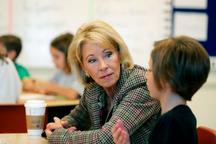Betsy DeVos in a grey-black checked blazer talking to a child while sitting