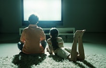 A man and his daughter sitting on the carpet in front of their large tv as it shines its light on th...