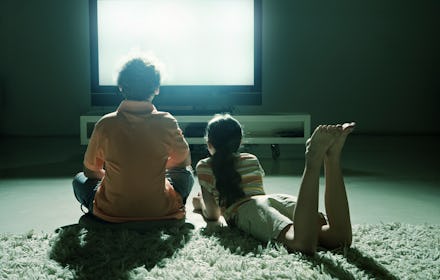 A man and his daughter sitting on the carpet in front of their large tv as it shines its light on th...