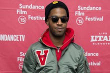 Kid Cudi posing in a green jacket, black cap, and black sunglasses