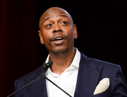 Dave Chapelle in a black suit, and white shirt talking into a microphone