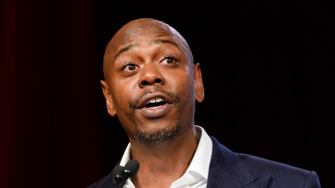 Dave Chapelle in a black suit, and white shirt talking into a microphone