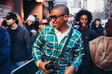 Mikey Alfred in a white shirt, a green-yellow checked jacket, and sunglasses walking through a crowd...