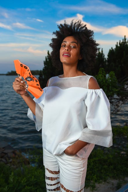 Solange Knowles in a white top and white trousers holding a small orange fan next to a lake