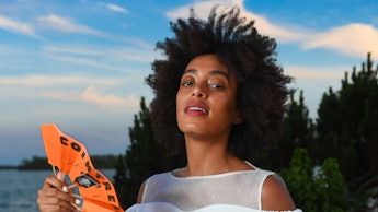 Solange Knowles in a white top and white trousers holding a small orange fan next to a lake