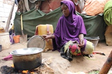 A woman in somalia crouched on the ground cooking a meal in the pot next to her