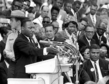 A black and white photo of Martin Luther King giving a speech surrounded by people and a police offi...