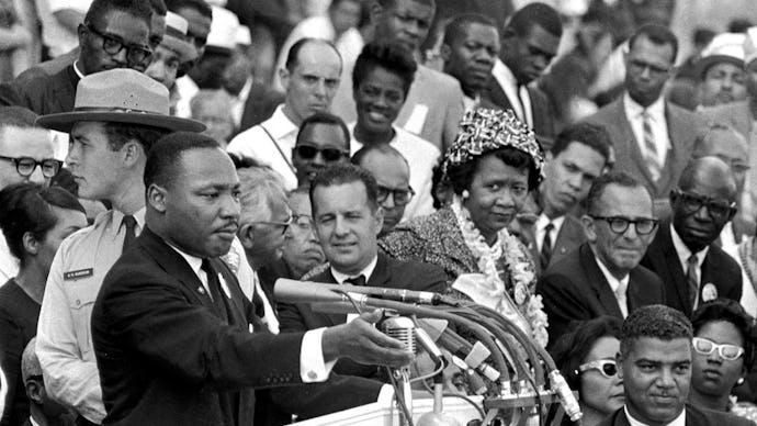 A black and white photo of Martin Luther King giving a speech surrounded by people and a police offi...