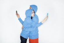 A couple in matching blue jackets hugging each other but looking at their phones at the same time