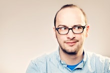 Josh Gondelman posing in glasses and a polo shirt over a blue shirt