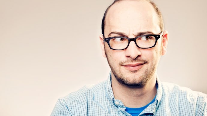 Josh Gondelman posing in glasses and a polo shirt over a blue shirt