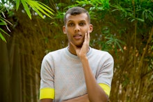 Stromae in a grey and yellow shirt with his hand on his left cheek