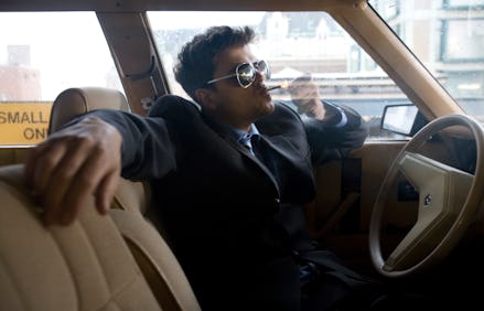 A man in a black jacket and sunglasses with his arm leaned against the car seat, smoking marijuana i...