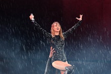 Taylor Swift on stage being picked up by someone, wearing a bedazzled black bodysuit as it rains