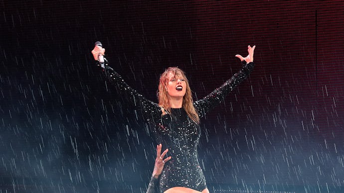 Taylor Swift on stage being picked up by someone, wearing a bedazzled black bodysuit as it rains