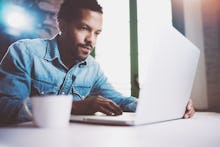 A man in a blue shirt who got started as a freelancer sitting and typing on his laptop