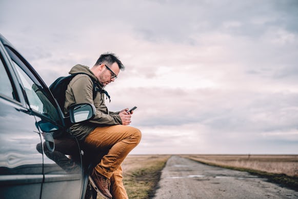 A man leaning onto his car and looking at his phone on the road during a solo trip across America