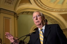 Mitch McConell in a black suit, a blue shirt, and a blue-yellow tie speaking into a microphone