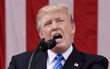 Donald Trump in a black suit, a white shirt, and a blue tie speaking into a microphone 