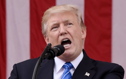 Donald Trump in a black suit, a white shirt, and a blue tie speaking into a microphone
