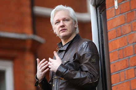 Julian Assange in a black leather jacket standing next to a red brick wall