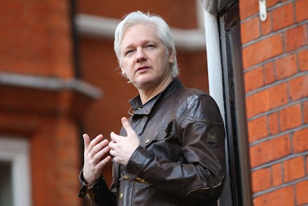 Julian Assange in a black leather jacket standing next to a red brick wall 