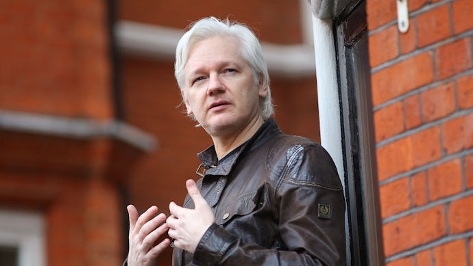 Julian Assange in a black leather jacket standing next to a red brick wall 