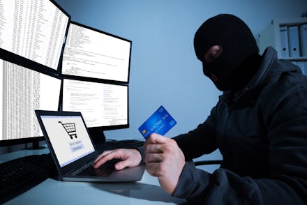 A man in a ski mask holding a credit card in front of an open laptop and four computer screens