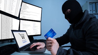 A man in a ski mask holding a credit card in front of an open laptop and four computer screens