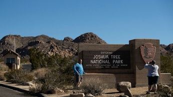 Two people stand in front of the sign for joshua tree national park and take a picture