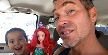 A dad sitting in a car next to his son, who is holding a 'Little Mermaid' doll
