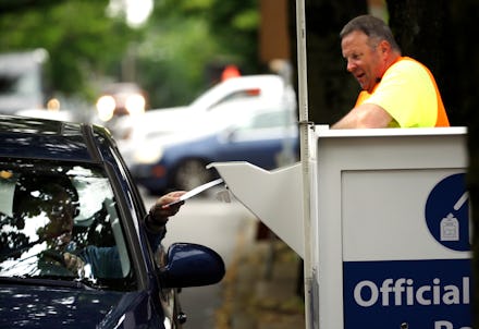 A man working a drive by voting booth and a person in a car handing over their ballot 