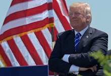 Donald trump with his arms crossed, a smirk on his face standing in front of an american flag