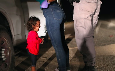 An immigrant child standing in front of a woman wearing jeans and purple rubber gloves, and a man in...